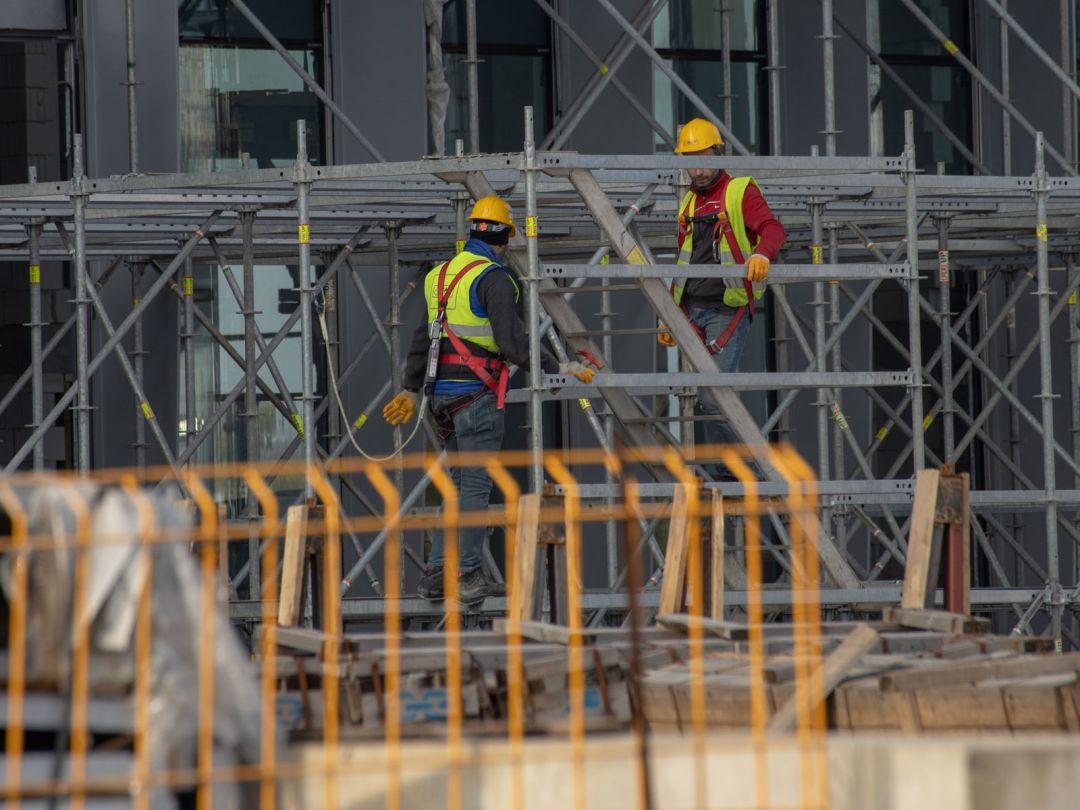 Zwei Gerüstbauer mit Helm, Warnweste und Auffanggurt bauen an einem Fassadengerüst aus Stahlrahmen